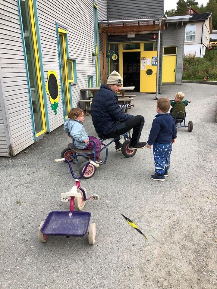 Halvor Egner Granerud sykler trehjulssykkel med et barn bakpå og prater med et annet barn som står foran han.