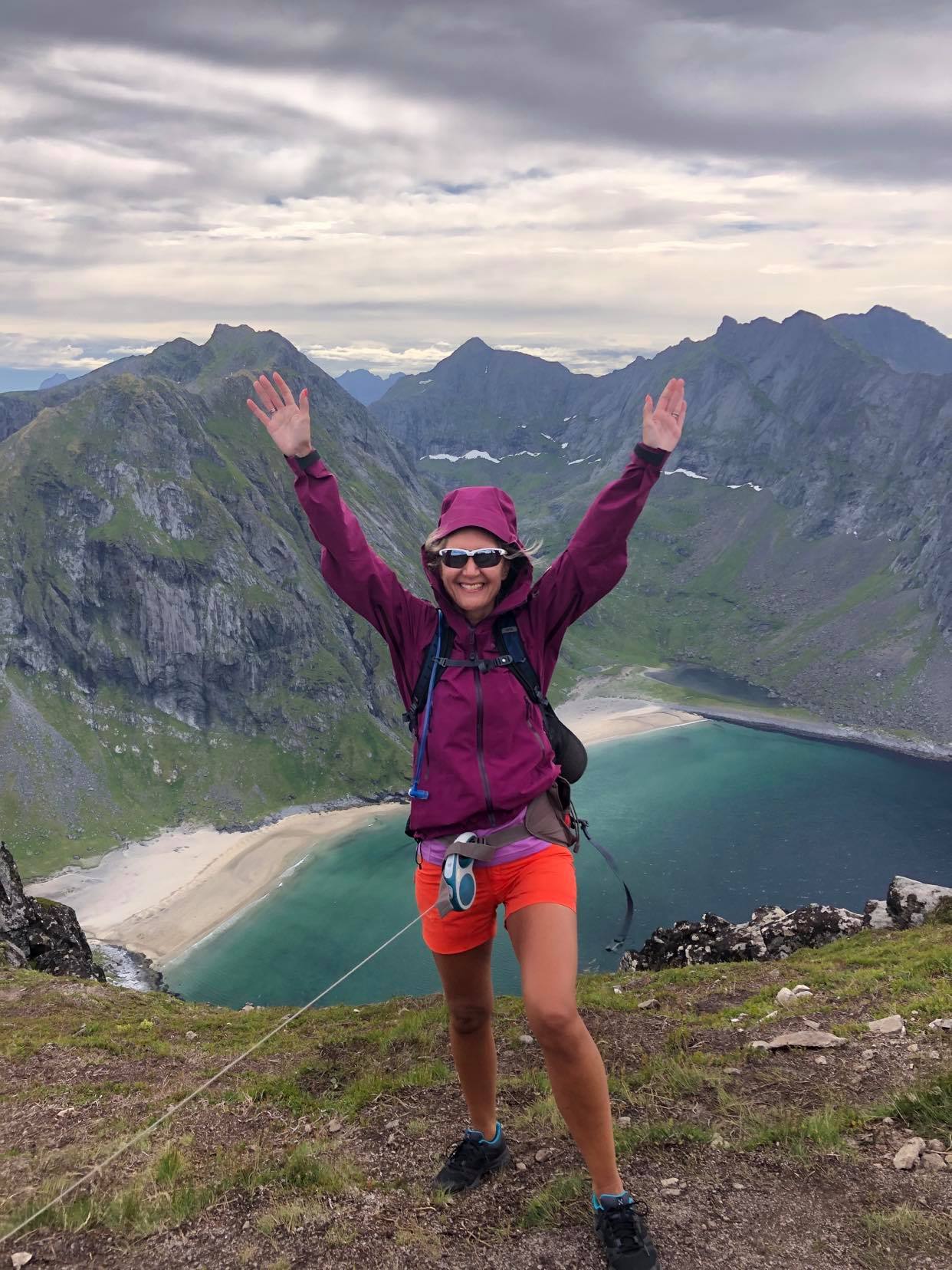 Hilde står i lilla jakke og shorts på toppen av et fjell i shorts og solbriller. I bakgrunnen er det storslått utsikt over fjell og strandlinje