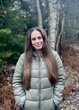 Lys ung dame med langt mørkt hår står med hendene i lommen på boblejakke langs en skogssti.