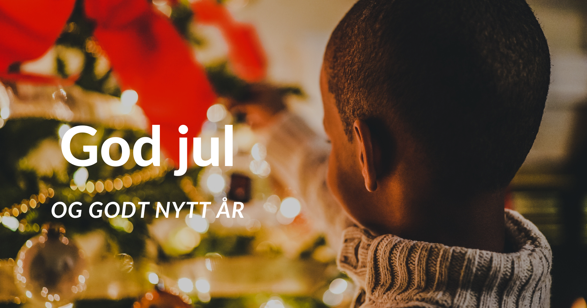 God jul i tekst over juletre som en melaninrik gutt med kort, svart hår står og pynter (med bakhodet mot kamera).